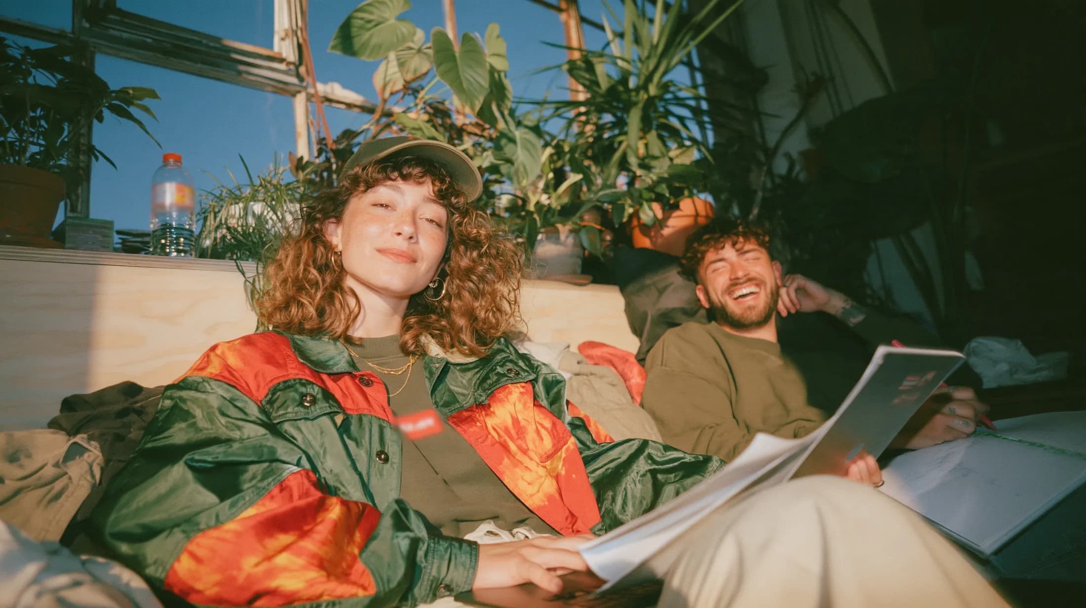 Two colleagues sitting relaxed by a sunny window full of plants, she smiles into the camera and he laughs over his notes.